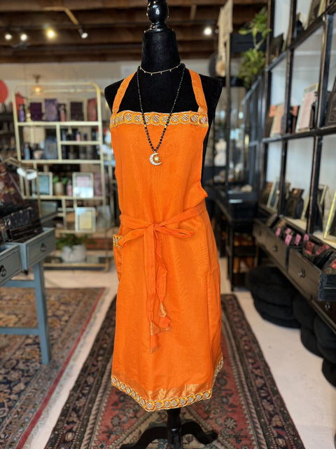 A mannequin showcases a vibrant orange apron from the Cool & Casual Handmade Sari Aprons collection, embellished with intricate hand-embroidered borders around the hem, pockets, neckline, and waist tie. Complementing the apron is a crescent moon pendant necklace. The cozy store background features shelves filled with plants and rugs from Casual Cool Aprons.