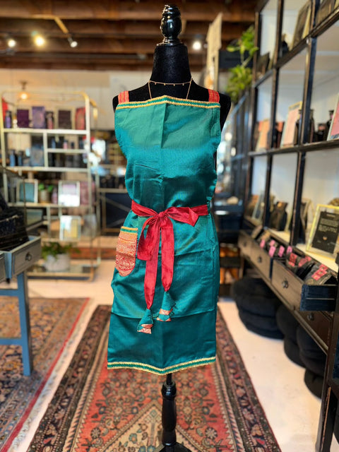 A mannequin dressed in a teal apron with a red belt and pocket is centered in a warmly lit boutique. The apron, from the Cool & Casual Handmade Sari Aprons collection, features intricate hand-embroidered details at the hem. The background showcases shelves with small items and books, along with a colorful, patterned rug on the floor.