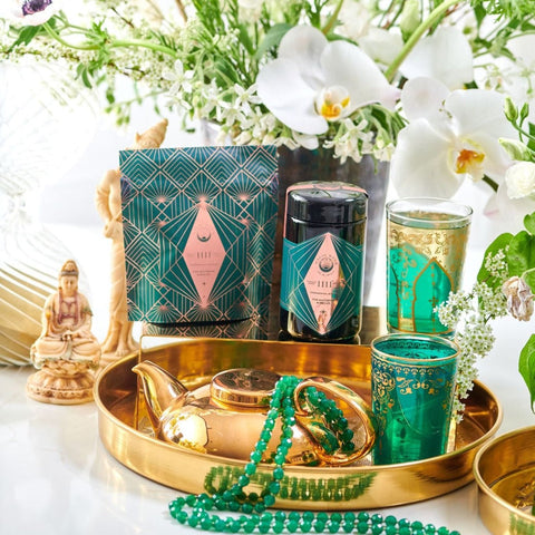 A luxurious setting features an elegant green and gold tea set on a gold tray, accompanied by a beaded green necklace, a small Buddha statue, and delicate white flowers in the background. The chic packaging suggests an artisanal product or gift set, perfect for enjoying Matcha 1111 : Highest Grade of Ceremonial Matcha by Magic Hour.