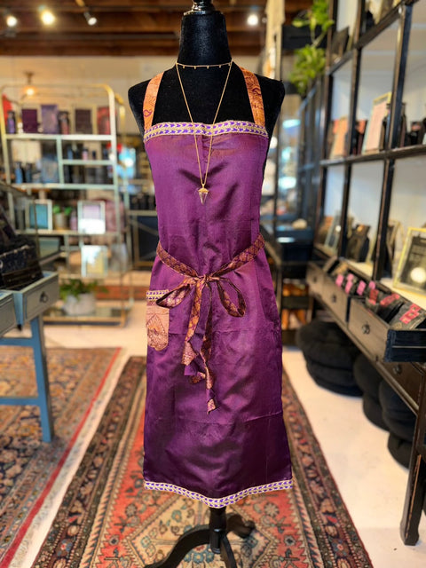 A mannequin displays the Cool & Casual Handmade Sari Apron, a deep purple apron with decorative trim and a front pocket, in a store setting. This unique apron is tied at the waist with a patterned sash and features exquisite hand-embroidered details. The store also offers various items on shelves and an eye-catching patterned rug enhances the floor.