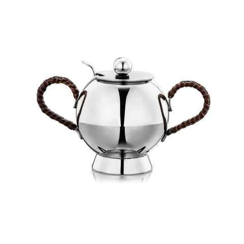 The Spheres Sugar Bowl - Wicker Handle is a polished stainless steel sugar bowl with two curved synthetic wicker handles wrapped in brown rattan. It features a round lid with a small knob on top and a slot for a spoon. The bowl has a reflective surface and stands on a short, wide base.