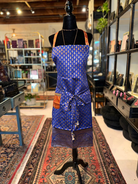 The Elevate Everyday Handmade Sari Apron, featuring a blue fabric with an intricate white pattern, is displayed on a black mannequin in a store. This luxury apron includes a pocket on the left side and is tied at the waist with a matching sash. The store, adorned with colorful rugs and shelves filled with various items in the background, showcases hand-embellished designs.