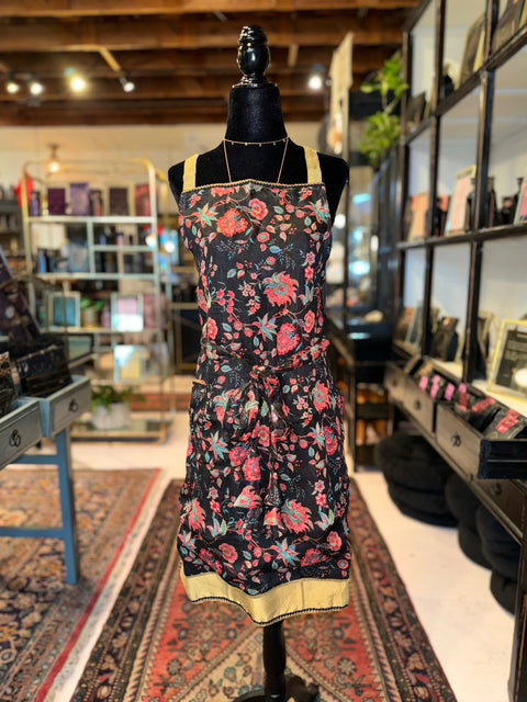 A black mannequin displays the Elevate Everyday Handmade Sari Apron, adorned with hand-embellished floral-patterned designs featuring pink and red roses and yellow trim. It stands in a rustic store with wooden shelves filled with various items. The background includes rugs, plants, and other merchandise, creating a cozy ambiance.