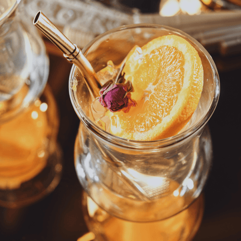 A refreshing glass of iced tea, crafted with loose tea and elegantly garnished with a slice of orange and a delicate rose bud. The drink is highlighted by the use of the Sips Tea Straw & Strainer Bombilla Case Packs from Alibaba, and is set aglow by warm, ambient light.