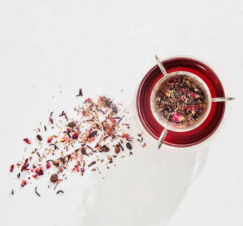 A glass cup filled with Immune Boosting Teas Sampler Set by Magic Hour and a tea strainer containing loose leaf tea rest on a white surface. A small pile of scattered tea leaves is visible beside the cup. The organic tea has a deep red color, and the scene is brightly lit.