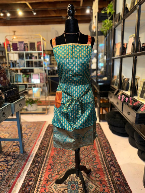 A mannequin draped in an Elevate Everyday Handmade Sari Apron, featuring luxurious green fabric with orange and gold accents, is displayed in a cozy, eclectic shop filled with books, ornaments, and rugs. The background has shelves lined with various items and a detailed woven print rug beneath the mannequin.