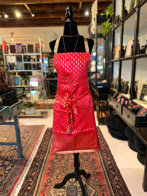 A mannequin dressed in the Elevate Everyday Handmade Sari Apron, featuring exquisite hand-embellished designs, stands on an intricate rug in a well-lit boutique. Shelves filled with various items line the walls, and the wooden ceiling adds a rustic touch to the cozy, eclectic shop atmosphere.