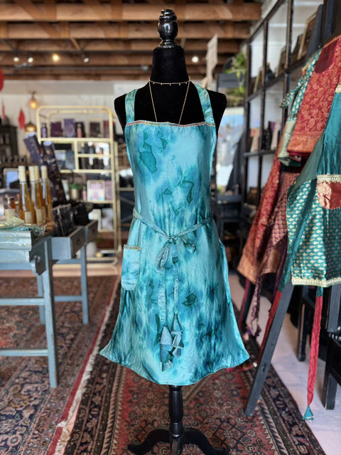 In a cozy, eclectic shop with wooden beams and decorative rugs, a Elevate Everyday Handmade Sari Apron is displayed on a black mannequin. The background includes shelves adorned with various items and colorful fabrics.