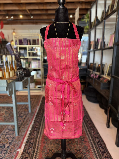 An Elevate Everyday Handmade Sari Apron, adorned in magenta with intricate golden patterns, is displayed on a black mannequin in a cozy, rustic shop. The store features bookshelves filled with various items, a patterned rug, and bottles on a table in the background, highlighting the luxury aprons and their exquisite hand-embellished designs.