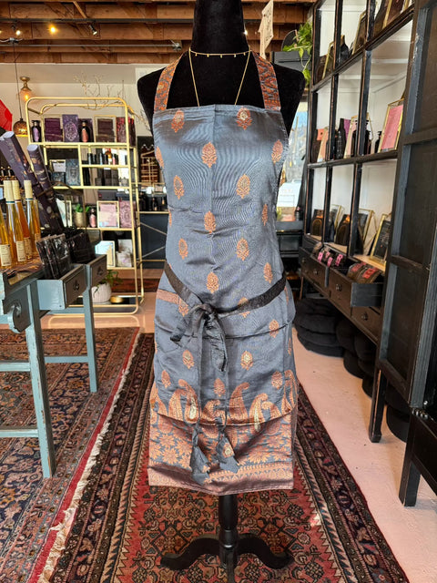 In the store, a display mannequin is adorned with an Elevate Everyday Handmade Sari Apron, featuring intricate patterns in shades of blue and copper. Behind the mannequin, shelves filled with various items like bottles and books create a cozy ambiance, while patterned rugs cover the floor.