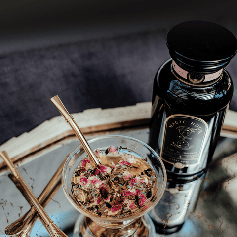 A jar labeled "Magic Hour" stands next to a glass filled with a mixture of herbs and dried rose petals, complemented by an Alibaba Sips Tea Straw & Strainer Bombilla resting inside. Both items are set on a reflective surface, beautifully capturing the light.