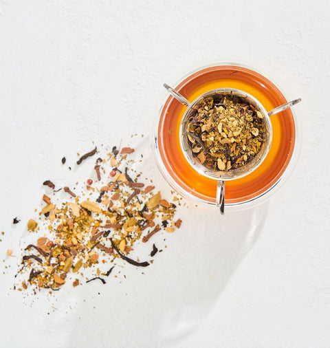 A glass teapot filled with orange-hued Magic Hour Immune Boosting Teas Sampler Set is photographed from above. Loose Leaf Tea and herbs are scattered beside the pot on a white surface, creating a colorful mix of ingredients. The image is bright with natural light casting soft shadows.