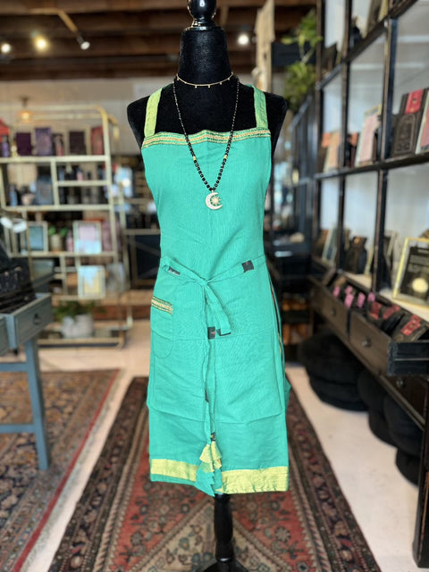 A mannequin dressed in a green apron with yellow trim from our Cool & Casual Handmade Sari Aprons collection is displayed in the center of the store. The apron features pockets and a tie around the waist. In the background, shelves and tables filled with books and various items are visible, along with a large woven rug on the floor.