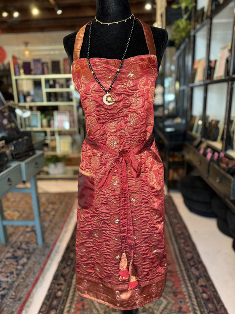 A mannequin displays the "Elevate Everyday Handmade Sari Apron," a luxurious piece featuring intricate, hand-embellished red and gold brocade designs with tie straps. The apron is complemented by a black beaded necklace adorned with a crescent moon pendant. The background reveals a store interior filled with an array of items on neatly arranged shelves.