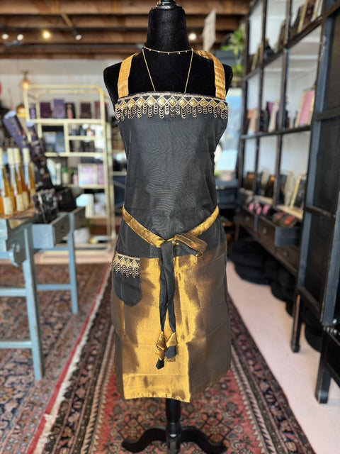 A black mannequin in a store showcases the Elevate Everyday Handmade Sari Apron, featuring intricate black and gold woven prints. In the background, shelves are stocked with various items, including bottles, books, and decor, alongside a large patterned rug on the floor.