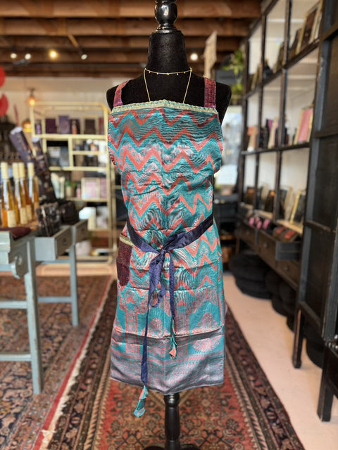 In a cozy, well-lit store, a mannequin is showcased wearing the "Elevate Everyday Handmade Sari Apron," adorned with colorful, intricately patterned zigzag designs in various shades of blue, green, pink, and purple. This luxury apron features hand-embellished details and is elegantly tied at the waist with a ribbon. Shelves filled with various items create an inviting backdrop.