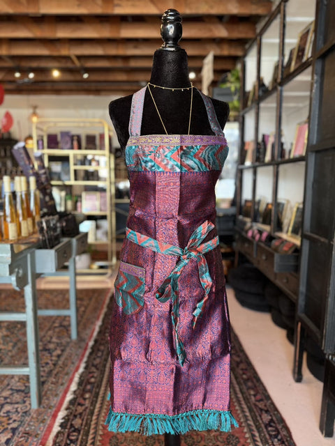 A mannequin showcases the "Elevate Everyday Handmade Sari Apron," featuring an intricately designed luxury apron with a purple background, turquoise trim, and a multicolored geometric pattern. This hand-embellished apron includes a front pocket and a tied waistband. The display is set in a rustic shop surrounded by shelves and various items in the background.