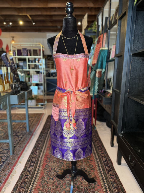 A mannequin draped in the Elevate Everyday Handmade Sari Apron, featuring a luxurious blend of orange and purple hues with gold floral patterns, stands in a boutique. The hand-embellished design pairs perfectly with the layered necklaces. The store has patterned rugs and shelves filled with various colorful fabrics.