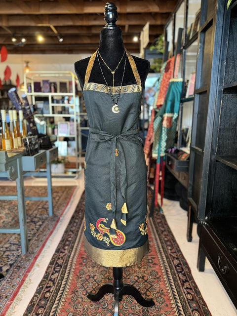 A black mannequin is dressed in the Elevate Everyday Handmade Sari Apron, featuring luxurious gold and colorful embroidery around the neckline and hem. The apron is cinched at the waist with a matching sash. The mannequin stands in a cozy boutique, surrounded by shelves and display tables showcasing hand-embellished designs.