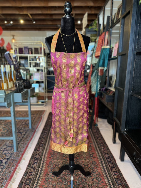 A black mannequin is adorned with a Fancy & Adorned Handmade Sari Apron that showcases a vibrant gold and pink floral design. This luxurious apron features a golden yellow hem and is displayed in a beautifully decorated store with shelves containing folded fabrics and other items in the background.