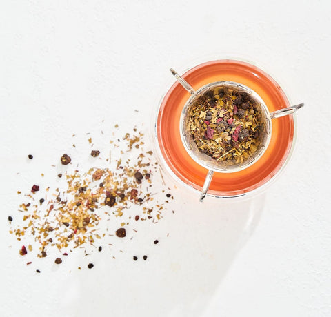 A glass mug filled with organic tea sits on a white surface. Above it, a metal tea strainer holding loose leaf tea is placed in the rim of the mug. To the left, some loose tea leaves are scattered on the surface, enhancing this Magic Hour Immune Boosting Teas Sampler Set moment.