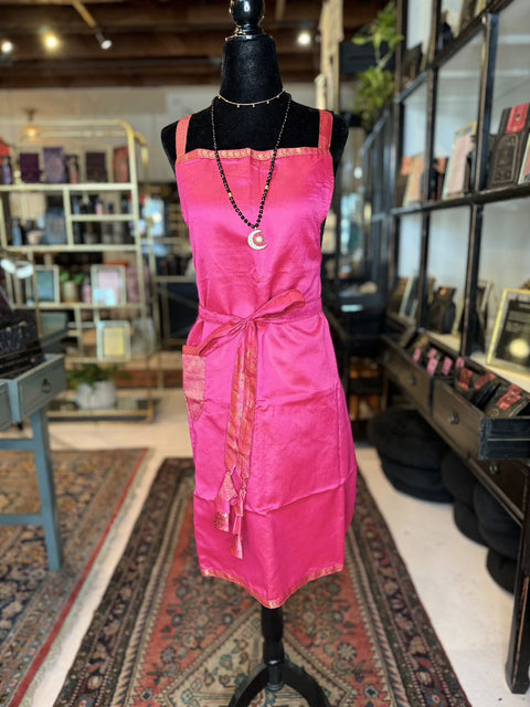 In a bookstore, a mannequin is adorned with a bright pink apron from the Cool & Casual Handmade Sari Aprons collection. The apron features hand-embroidered details and is tied at the waist with a sash, accented by gold trim at the top. Shelves brimming with books and a rustic interior add to the charming backdrop.