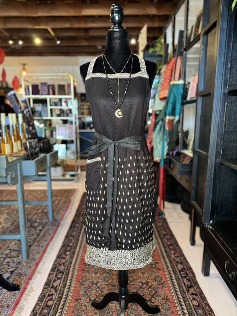 A black dress with a white pattern is displayed on a mannequin in a boutique. The dress features a halter neckline and a decorative white border at the hem. Nearby, Fancy & Adorned Handmade Sari Aprons hang elegantly amidst the rugs, tables, and shelves filled with various items and colorful fabrics.