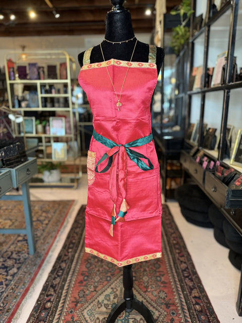 A mannequin displays one of our unique Cool & Casual Handmade Sari Aprons, featuring a red apron with hand-embroidered details, green ties, and gold trim, over a patterned blue and red rug. The apron also includes a pocket on the right side and is adorned with a necklace featuring a pendant draped over it. Shelves with various items are visible in the background.