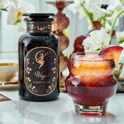 A glass of iced plum-colored drink garnished with a plum slice sits next to a black bottle labeled "Virgo Tea of Virtue, Wit & Meticulous Magic" decorated with gold celestial designs. The background features a teacup, white flowers, and decorative vases, evoking the soothing essence of Magic Hour.