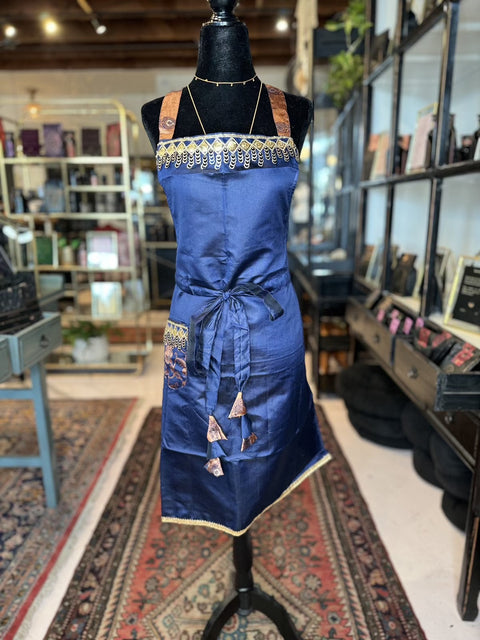 A mannequin in a store showcases one of our Cool & Casual Handmade Sari Aprons, featuring a blue apron with intricate gold and hand-embroidered details. The apron boasts brown and gold straps and a front pocket adorned with a detailed design. Rugs, shelves, and various items create an inviting backdrop in the store.