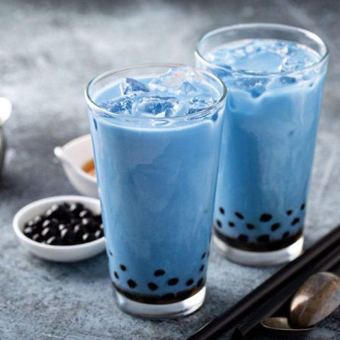 Two clear glasses filled with vibrant blue bubble tea, topped with ice cubes. The drinks contain dark Boba : Tapioca Pearls settled at the bottom. The background includes small white bowls with additional ingredients and utensils, setting the scene like a Magic Hour tea party full of delightful surprises.