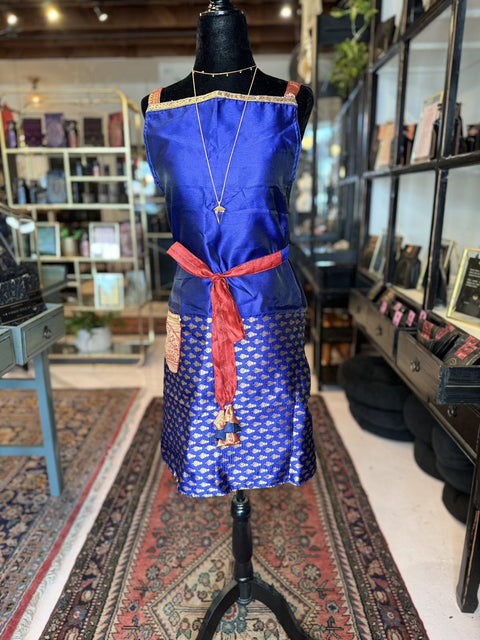 A mannequin dressed in a Cool & Casual Handmade Sari Apron, featuring hand-embroidered details and a red belt, is displayed in a stylish store. Behind it, shelves hold various products, including bags and other accessories. The store has a cozy ambiance with decorative rugs on the floor.