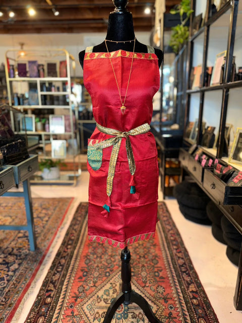 A mannequin dressed in a Cool & Casual Handmade Sari Apron featuring red fabric with gold trim and hand-embroidered details stands on a rug in a boutique. The apron is accessorized with a long necklace. Shelves filled with books and trinkets line the background, accompanied by a window and various framed items, adding charm to the setting.