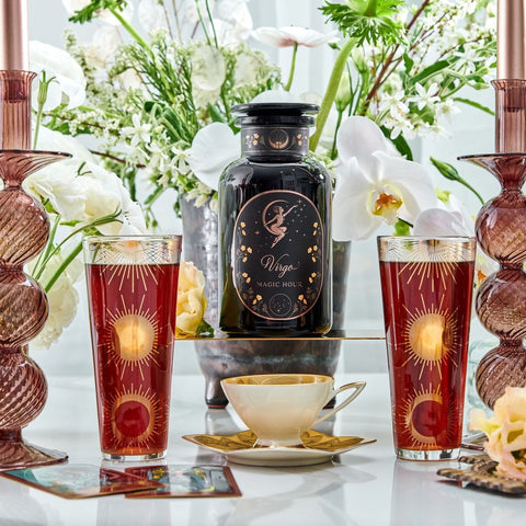 A decorative scene with a large black apothecary jar labeled "Virgo Tea of Virtue, Wit & Meticulous Magic" surrounded by white flowers. Two tall pinkish glasses with sun designs and a teacup of Magic Hour tea are placed on the table. Elegant candle holders and floral arrangements complete the setting.