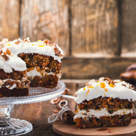 A single slice of Year of the Rabbit Carrot Cake with cream cheese frosting and chopped nuts sits on a wooden board, accompanied by a steaming cup of herbal tea. The rest of the cake is on a glass cake stand, against a rustic wooden background.