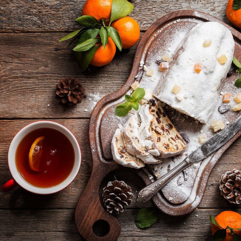 A wooden table displays a sliced stollen topped with powdered sugar and garnished with peppermint leaves and candied fruits on a wooden cutting board. Pinecones, tangerines with leaves, and a red cup of Sagittarius Tea of Good Fortune & Abundance by Magic Hour with a lemon slice are arranged around it.