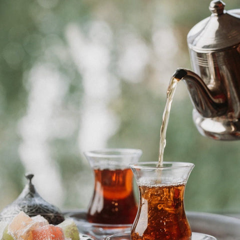 A close-up of two traditional glass cups filled with Magic Hour's Tea of The Rising Sun : Japanese Breakfast on a tray. A silver teapot is pouring tea into one of the cups. In the foreground, there are pieces of colorful Turkish delight. The background is blurred, showing green foliage.