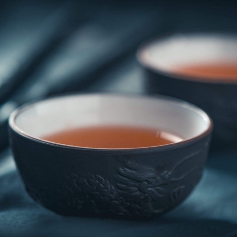 Close-up of a dark ceramic bowl filled with a warm brown liquid, likely Ti Quan Yin Oolong: Tea of Patient Compassion from Magic Hour, set against a blurred dark background. Another similar bowl is partially visible in the distance, suggesting a serene, contemplative setting that highlights the wellness benefits of this ancient brew.