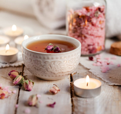 A delicate white ceramic cup filled with herbal tea, garnished with dried rosebuds, sits on a whitewashed wooden surface. Surrounding the cup are lit tea candles, scattered dried rose petals, and a glass jar containing Pink Tourmaline Bath Soak.