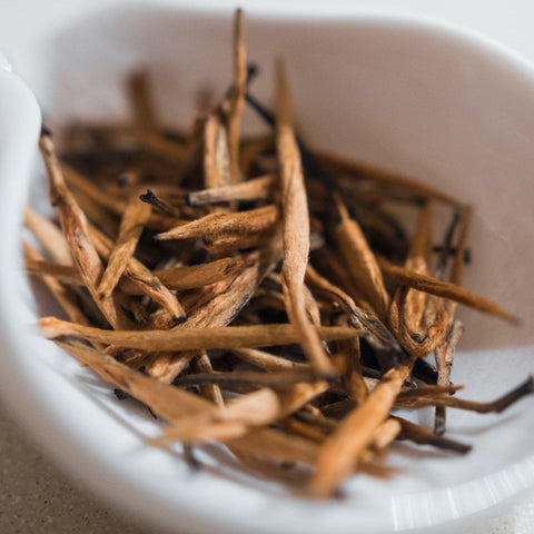 A white bowl filled with thin, dried brown tea leaves, arranged in a casual, slightly intertwined manner. This Organic Golden Monkey Tip Yunnan Black Tea from Magic Hour promises a rich infusion. The blurred background highlights the texture and details of these organic tea leaves.