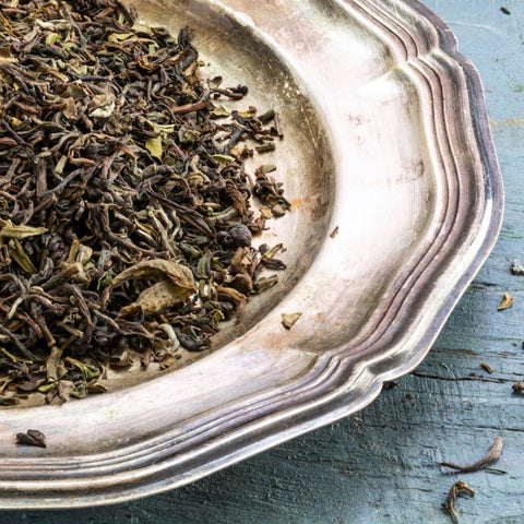 A close-up of a silver plate holding a pile of dried loose leaf tea from the Magic Hour Goomtee Estate Darjeeling. The leaves are a mix of dark browns and greens, indicating various stages of oxidation. The plate rests on a weathered, blue-painted wooden surface.