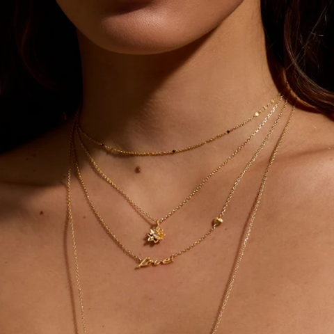 Close-up of a person's neck adorned with the "Adorned with Love Gold Necklace," an 18kt gold plated piece that adds elegance to a personalized layered look. The necklace features various pendants, including one in delicate script spelling "love," a floral design, and a small heart. The lighting casts soft shadows, highlighting the jewelry beautifully.