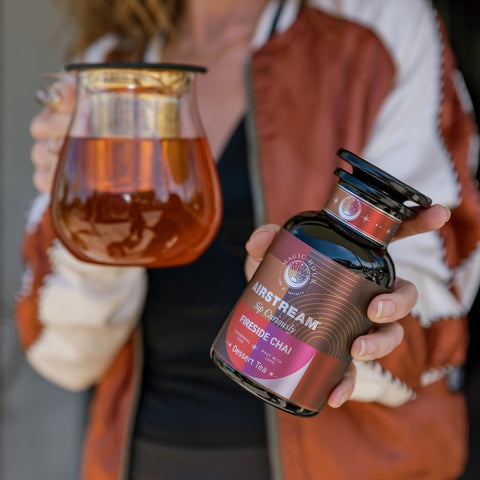 A person wearing a red and white jacket holds a glass pitcher of tea in one hand and a container labeled "Magic Hour - Fireside Chai: Warming Rooibos Blend for Creating Connection" in the other. The label, which features a red and black design, highlights that it's a caffeine-free spiced herbal chai packed with antioxidants including vitamin C.