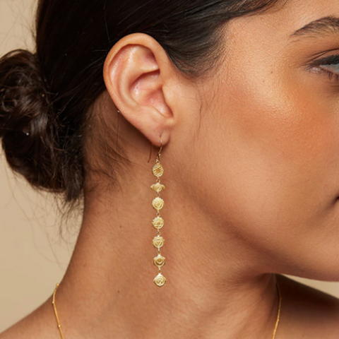 A woman with her hair in a bun is wearing the Aligned in Harmony Earrings, which are long, 18kt gold-plated drop earrings adorned with multiple small, intricate charms. She has warm-toned, glowing skin and the background is a soft beige color, creating a harmonious balance in the image.