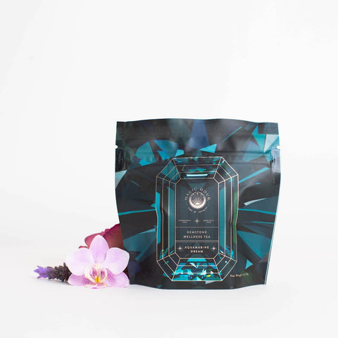 A pouch of "Magic Hour Aquamarine Dream - Soothing Herbal Ayurvedic Adrenal Tonic" stands against a white background. The packaging is designed with a gemstone graphic, featuring teal and black colors. In front of the pouch, there are two delicate pink orchids and a small purple flower, showcasing the allure of this organic loose leaf tea.