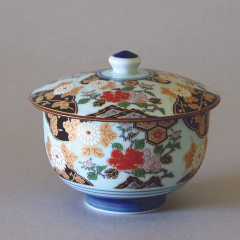 The Eastern Elm Arita Peony Flower Tea Cup with Lid is a decorative piece featuring intricate floral patterns in red, white, pink, and gold against a black background. The cup includes a blue base and a small knob on top of the lid, showcasing exquisite Japanese craftsmanship. The background is plain and light-colored.