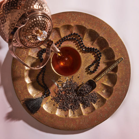 A copper teapot is pouring Magic Hour Assam Black Tea into a glass teacup placed on an ornate, round, golden tray. Next to the teacup, there is a pile of loose tea leaves known for their malty flavor and full-bodied character, a metal scoop, and a string of black beads with a tassel. The background is white with a soft shadow.