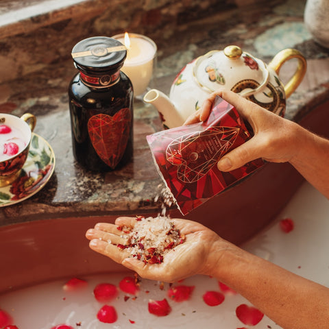 A pair of hands pours Pink Tourmaline Bath Soak from a red, heart-themed pack into one palm over a bathtub adorned with rose petals. Nearby, a black jar of organic tea seed oil and a decorative teapot with cups are placed on the edge of the tub, alongside a lit candle.