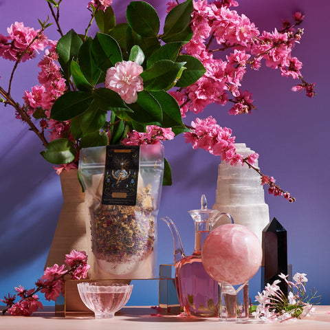 A stunning arrangement featuring the "High Priestess Bath Tea" from Magic Hour, pink cherry blossoms in a vase, a glass pitcher with pink liquid, crystal balls, and a black crystal obelisk, all set against a gradient purple and blue background that channels divine feminine energy.