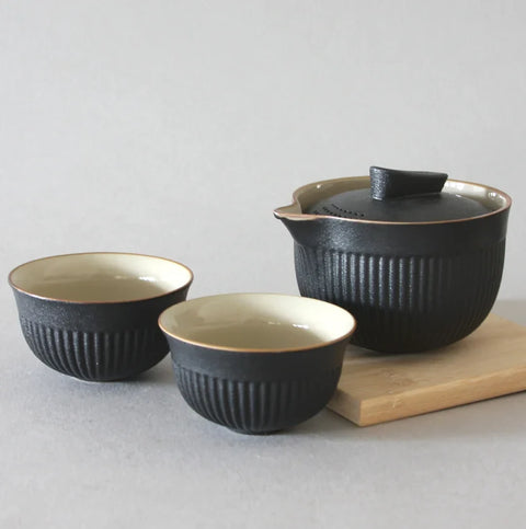 An Eastern Elm Black Travel Infuser Set sits elegantly, featuring a teapot and two matching cups with a textured, ribbed surface and a light-colored interior. The teapot rests on a wooden coaster against a plain gray background, embodying traditional craftsmanship perfect for any tea lover's collection.